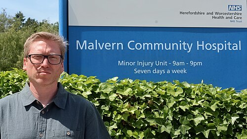 Cllr Dan, angry, next to Malvern NHS sign